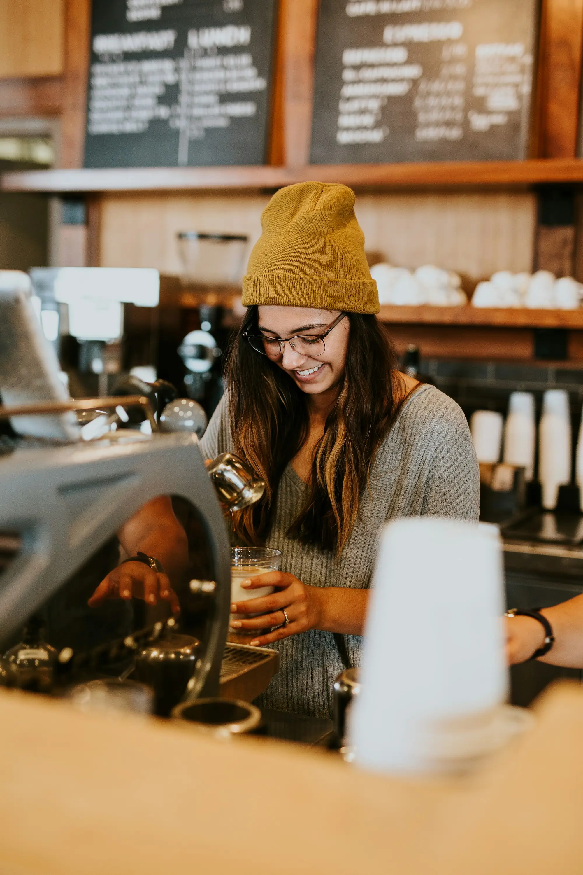 Cafe worker in a beanie at the espresso counter holding a glass.