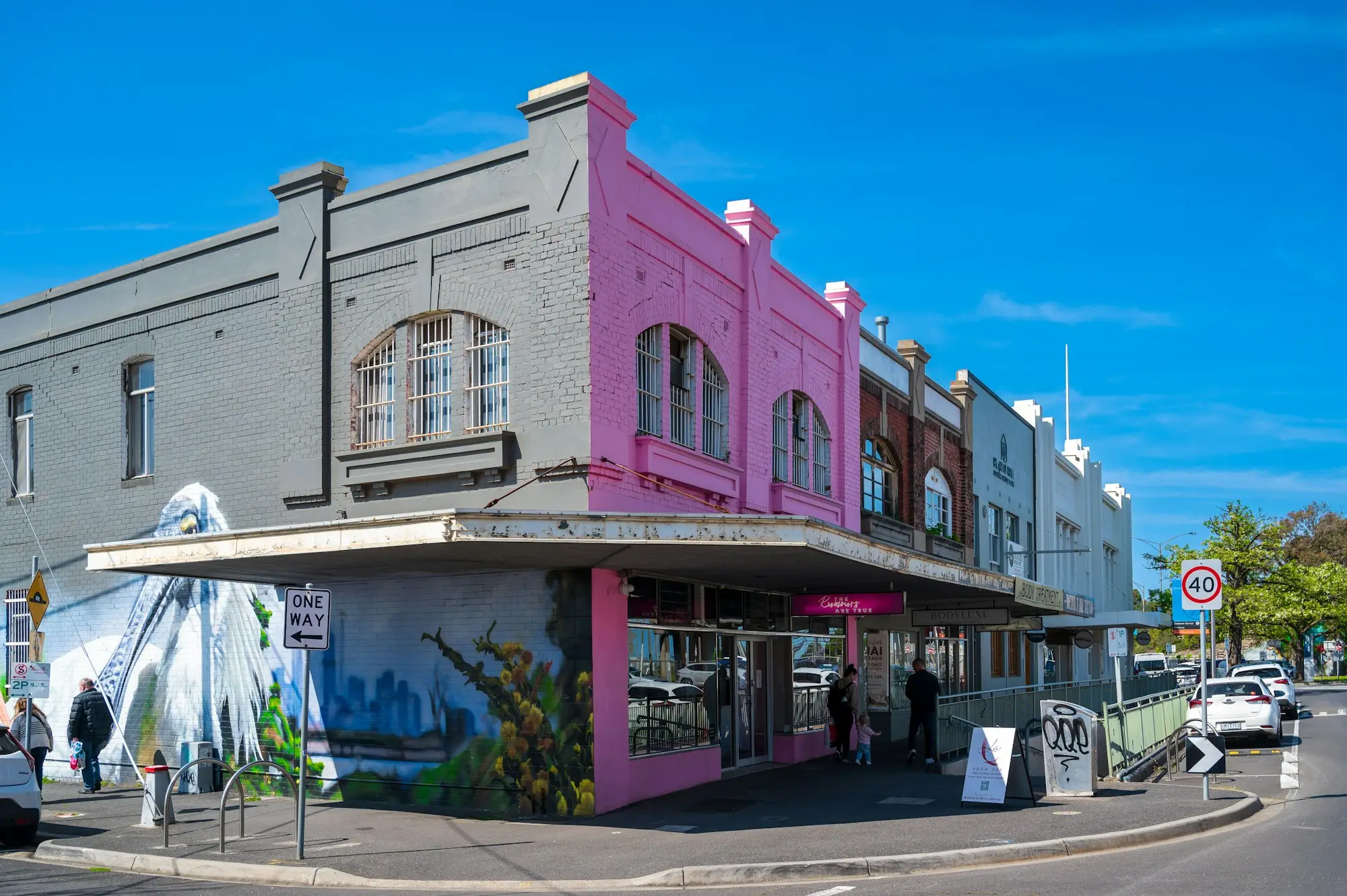 Two-storey corner building with shopfronts under a clear blue sky on a suburban street.