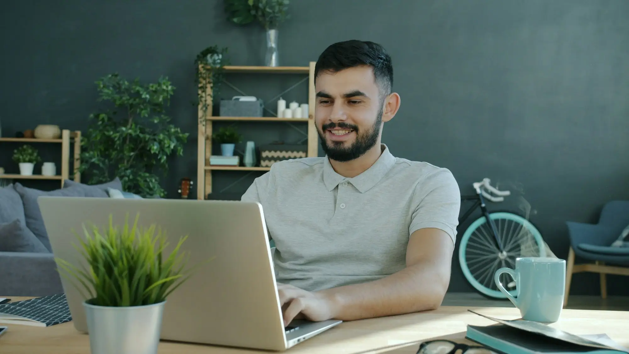 Business owner smiling at a laptop in a small home office with a plant and shelves.
