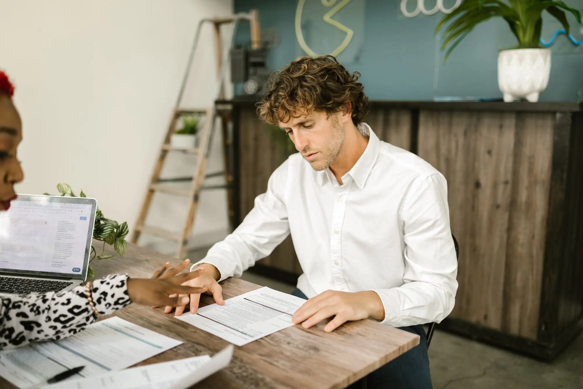 Business owner in a white shirt reading through printed bank statements at a timber table while an adviser points to a line